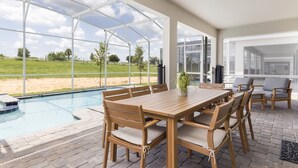 Een overdekt buitengebied met een zwembad, houten tafel en stoelen, en uitzicht op een grasveld.