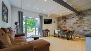 A modern living room with a leather sofa, a dining area with a table and chairs, a TV mounted on the brick wall, and a large window with curtains.