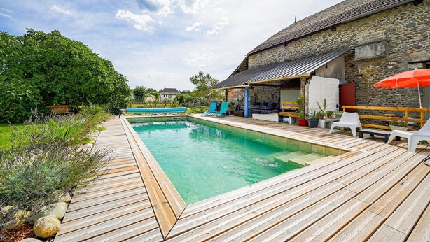 A wooden deck with a swimming pool, lounge chairs, and a stone building with a tiled roof.