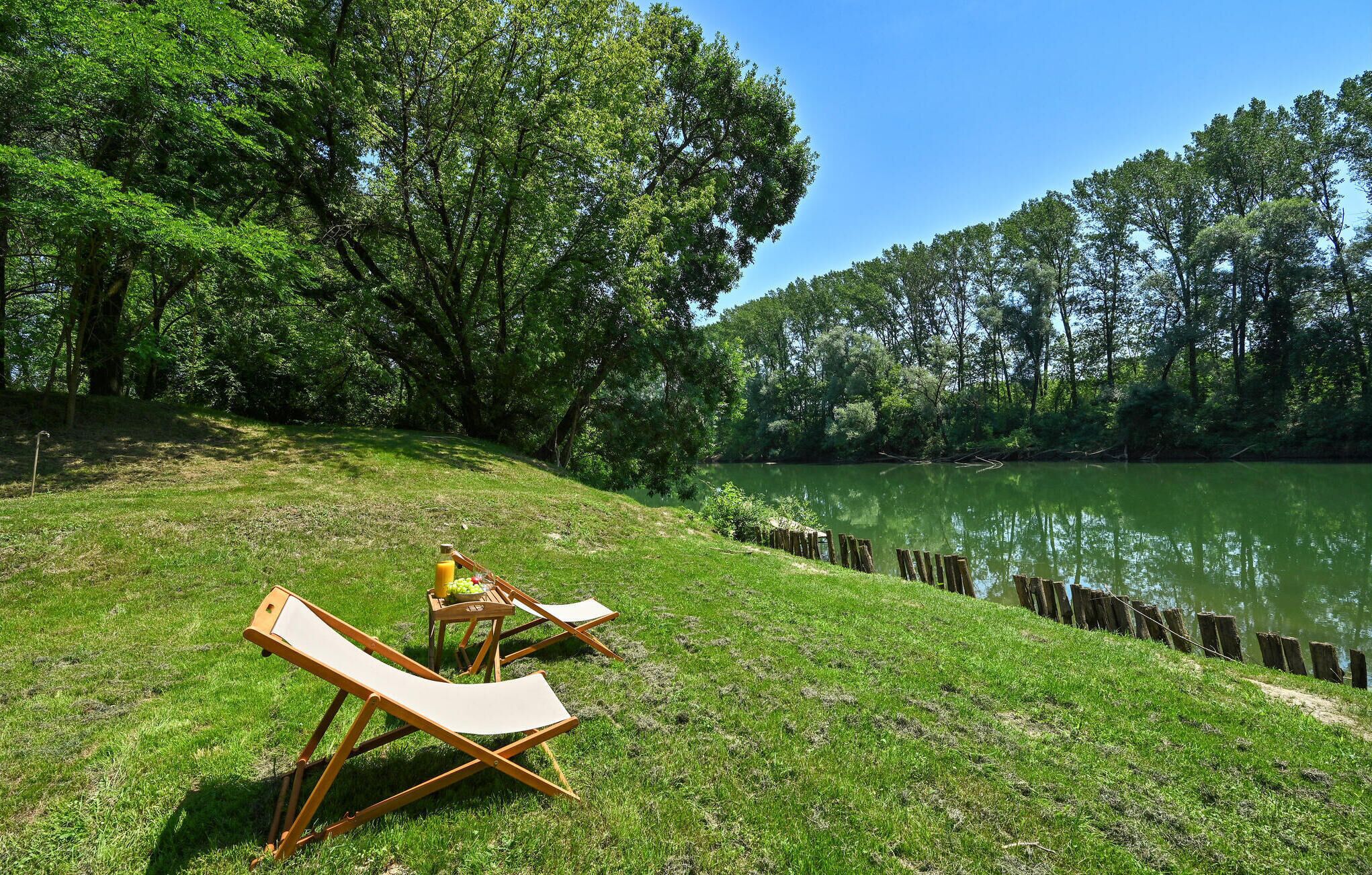 A grassy area with two lounge chairs and a small table with fruit and a bottle, near a calm lake surrounded by trees.