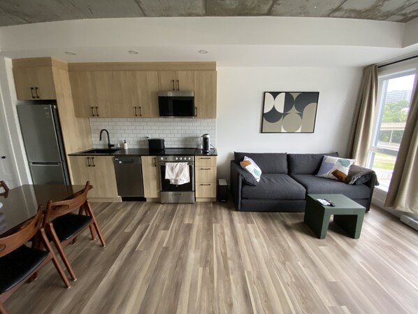A modern kitchen with wooden cabinets, a stainless steel refrigerator, and a black microwave. There is a dining table with chairs and a gray sofa with cushions. A painting is on the wall, and a window provides a view of the outdoors. - CONDO MID-CENTURY MODERN STYLE - POOL/TERRACE (Québec)
