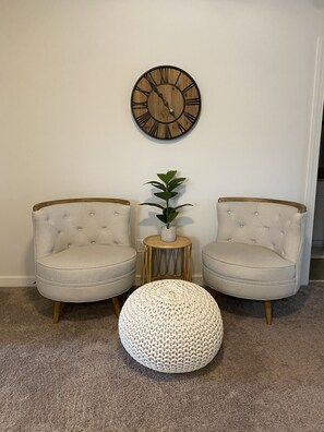 Une pièce comprenant deux fauteuils, un pouf rond en fibres tressées, une petite table d’appoint avec une plante et une horloge murale.