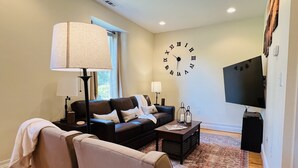 A living room with a sofa, a coffee table, a lamp, a clock, and a television. - Historic Schoolhouse on Winery Row (Purcellville)