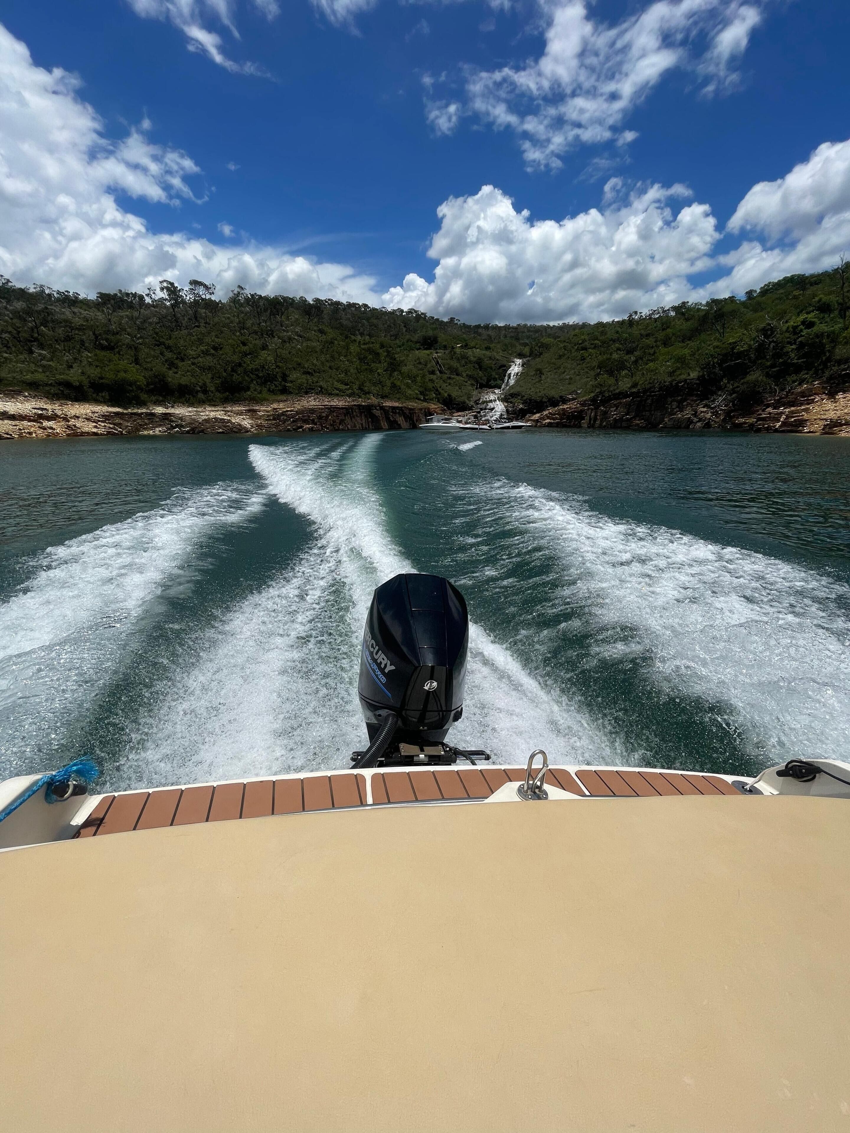 Um barco navegando por um rio, com uma cachoeira ao longe, rodeado por colinas florestadas.