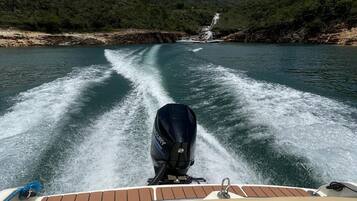 Um barco navegando por um rio, com uma cachoeira ao longe, rodeado por colinas florestadas.
