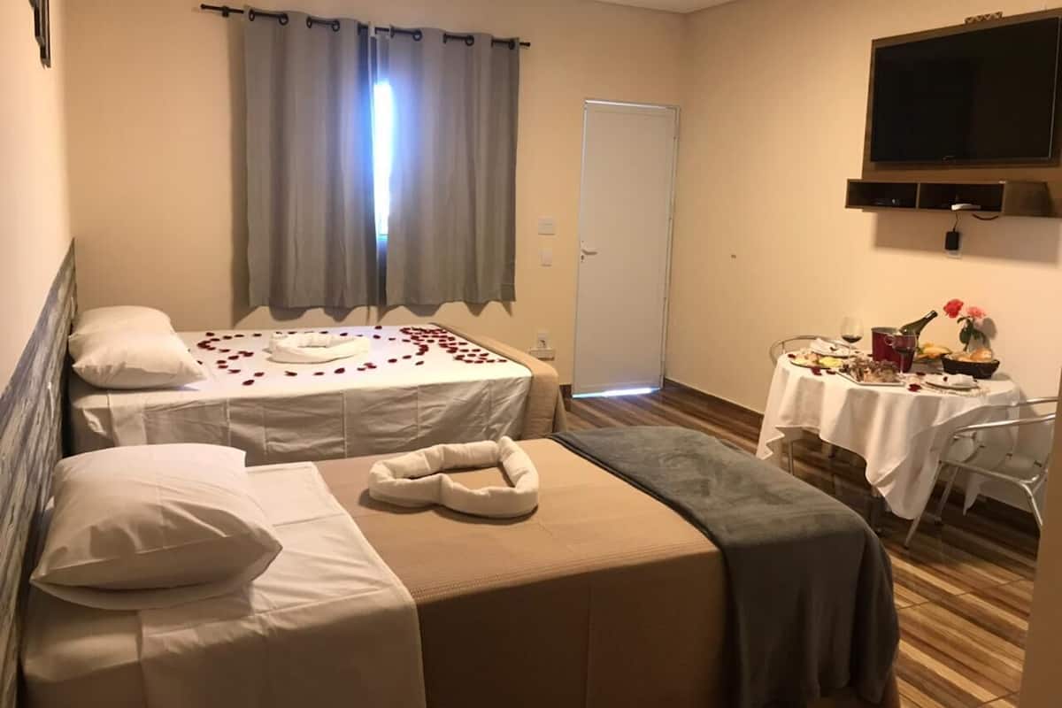 Ein Hotelzimmer mit zwei Betten, einem Esstisch mit Blumenstrauß und einem an der Wand montierten Fernseher.