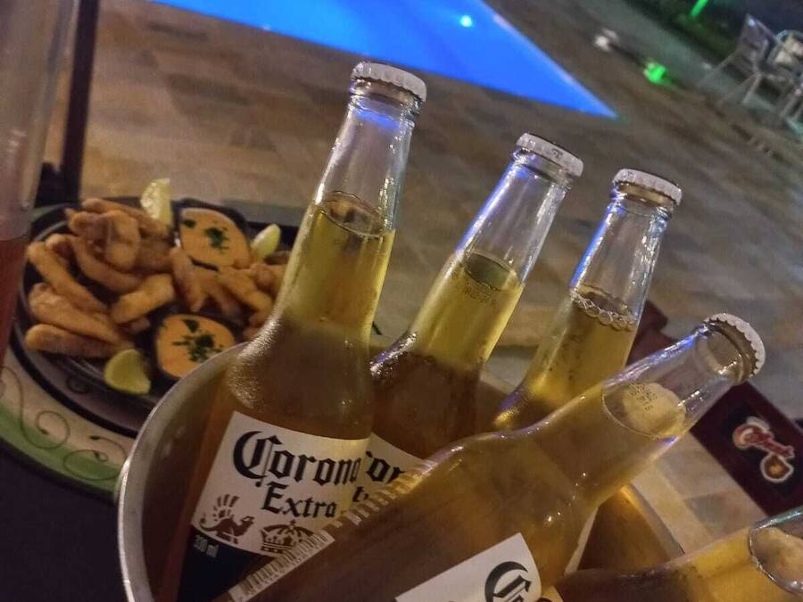 Um balde com quatro garrafas de cerveja Corona Extra, um prato de petiscos fritos e vista para a piscina com iluminação.