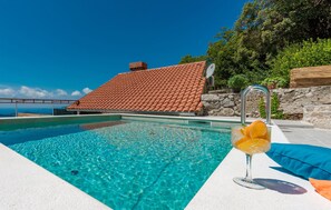 Une piscine d’un bleu éclatant, avec une maison au toit carrelé en arrière-plan, un verre de jus d’orange posé au bord de la piscine et un mur de pierre.