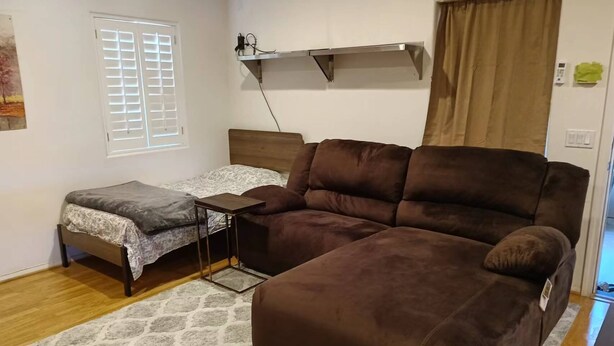 Un salon avec un canapé marron, un petit lit recouvert d’une courtepointe à motifs, une petite table, une fenêtre avec des stores et une étagère fixée au mur, équipée d’une prise électrique.