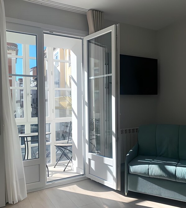 A room with a sliding glass door leading to a balcony, a green sofa, and a mounted flat-screen TV on the wall.