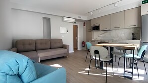 Een moderne woonkamer met een bank, een eettafel met stoelen, een keuken met kastjes en apparatuur, en een brandblusser.