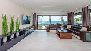 Ein modernes Wohnzimmer mit großem Flachbildfernseher, einer Couch, einem Couchtisch und Meerblick durch bodentiefe Fenster.