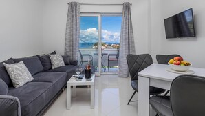 Ein modernes Wohnzimmer mit einem grauen Sofa, einem Esstisch mit Stühlen, einem an der Wand montierten Fernseher und einem Balkon mit Meerblick.