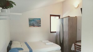 Een kleine, praktische kamer met een bed, bureau en stoel. Er hangt een schilderij aan de muur en er is een raam met gordijnen.