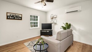 Una sala de estar moderna con ventilador de techo, televisor de pantalla plana sobre un soporte negro, sofá color beige, una pequeña mesa redonda con una planta y un cartel motivacional en la pared.