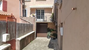 A narrow alleyway with a building on the left, a balcony with outdoor furniture, and a closed shutter door.