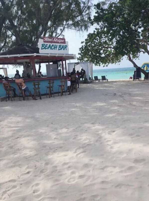 A beach bar with a sign reading "Red Stripe Beach Bar" and several patrons seated at the bar.
