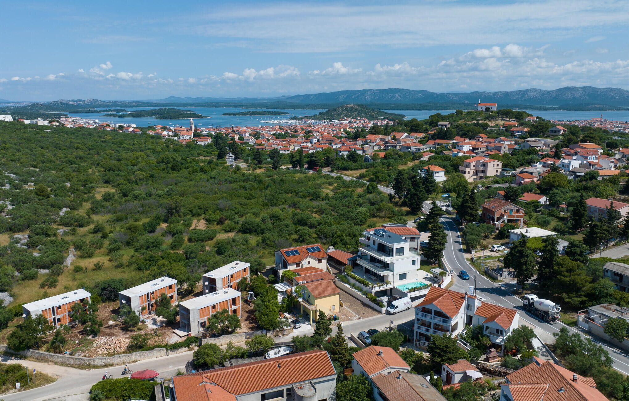Une ville côtière où se côtoient des immeubles résidentiels et des espaces verts, une route, et d’où l’on peut admirer la mer et les montagnes au loin.