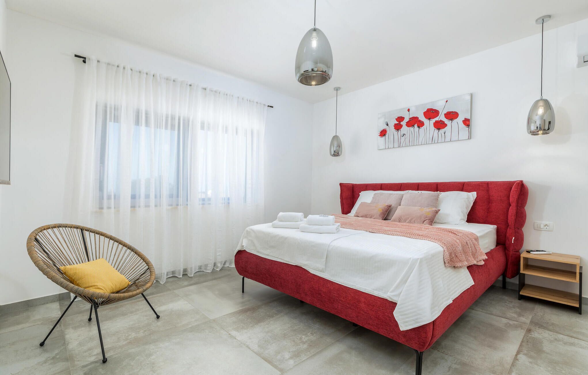 Ein modernes Schlafzimmer mit einem roten Bett, weißer Bettwäsche, einem Korbstuhl und einem großen Fenster mit leichtem Vorhang.