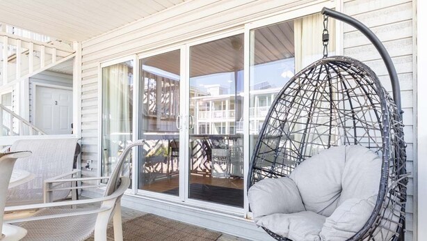 A hanging chair with a white cushion on a wooden deck, a white chair, and a table with a woven seat.