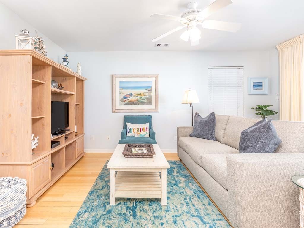 Een woonkamer met een houten televisiemeubel, een televisie, een blauw patroon tapijt, een beige bank en een plafondventilator.