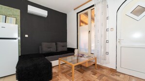 A compact living space with a sofa, a glass coffee table, a black furry ottoman, a white refrigerator, and a sliding glass door leading to a balcony.
