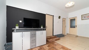 A modern living room with a flat-screen TV mounted on a black wall, a white cabinet with drawers, a wooden herringbone floor, and a door leading to another room.