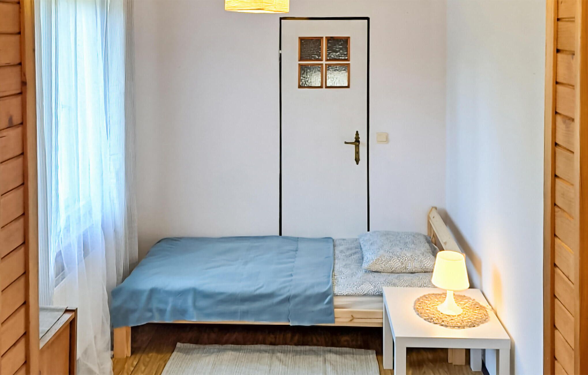 A small, neatly arranged bedroom with a single bed, a bedside table with a lamp, and a door with a window.