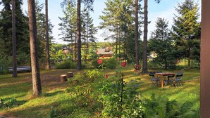 A forested area with a wooden table and chairs, a bench, and a house in the background.