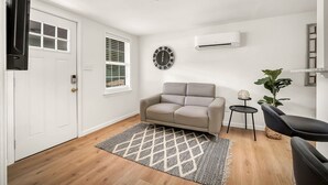 A modern living room with a beige sofa, a black armchair, a patterned rug, a wall clock, and an air conditioning unit. - One bedroom duplex in desirable Park Hill (Fort Smith)