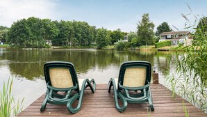 Een houten steiger met twee groene stoelen gericht op een rustig meer, omringd door weelderige begroeiing en huizen op de achtergrond.