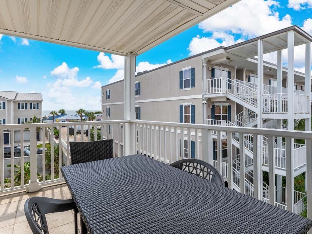 A covered outdoor area with a table and chairs, a white railing, and a view of a multi-story building with balconies.