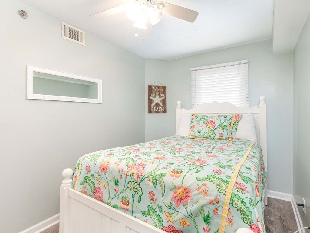 Une chambre à coucher comprenant un lit, un ventilateur de plafond, une fenêtre avec des stores, une étagère fixée au mur et un panneau décoratif.
