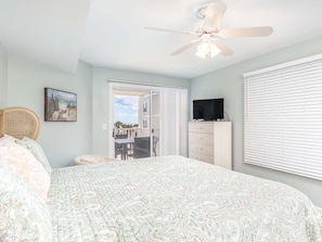 A bedroom with a bed, a ceiling fan, a TV, a dresser, a window with blinds, and a door leading to a balcony.