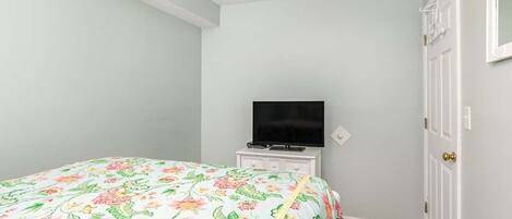 Une chambre à coucher avec une couvre-lit à motifs floraux, un téléviseur à écran plat posé sur une commode et une porte blanche.