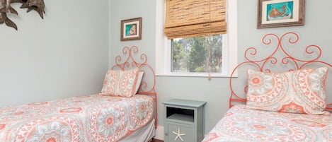 A bedroom with two beds, a small nightstand, a window with a bamboo blind, and wall art.