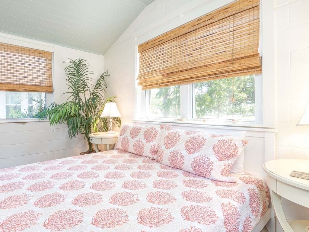 A bedroom with a bed, two windows with blinds, a bedside table, a lamp, and a potted plant.