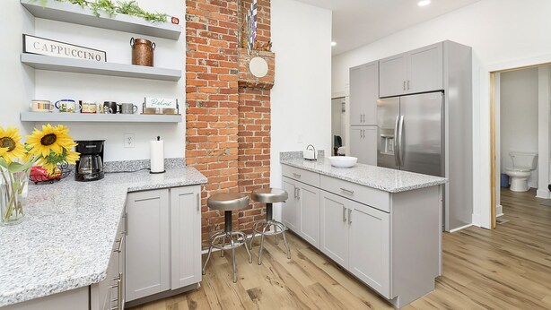 Une cuisine moderne avec des meubles blancs, un mur d’accent en briques, un réfrigérateur en acier inoxydable et un coin café.