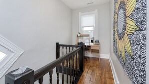 Un escalier avec une rampe en bois mène à une pièce où se trouvent un bureau et une chaise, une fenêtre avec des stores et un mur orné d’un motif.