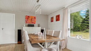 Un coin repas avec une table en bois, des chaises blanches et un lustre. Les murs sont ornés de peintures abstraites et une fenêtre offre une vue sur la verdure.