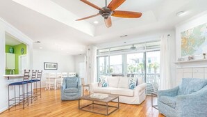 A spacious living room with a wooden floor, a ceiling fan, a white sofa, a blue armchair, and a dining area with a white table and chairs. - The Cool Coconut by Tybee Vacation Rentals (Tybee Island)