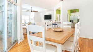 A dining area with a wooden table and white chairs, a ceiling fan, and a TV on the wall. - The Cool Coconut by Tybee Vacation Rentals (Tybee Island)