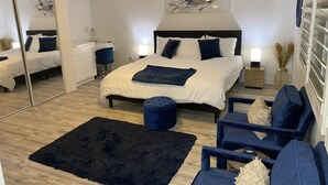 Un dormitorio moderno con una cama grande, un ventilador de techo, una mesita de noche con lámpara, un espejo y una silla azul oscuro.
