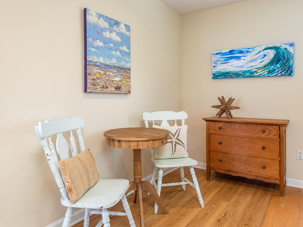 Een kamer met twee witte stoelen, een houten tafel, een houten dressoir en twee schilderijen met een strandthema aan de muur.