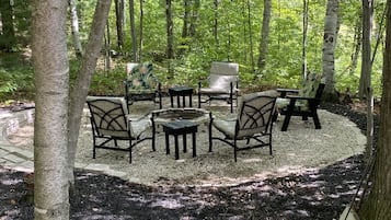 A forested area with a circular seating arrangement of chairs and tables, surrounded by trees and mulch.
