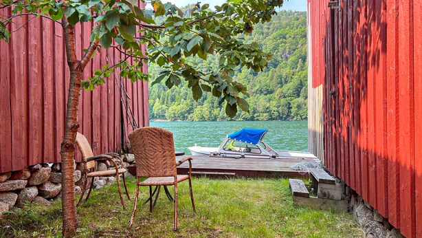 Eine Holzterrasse mit einem Boot und Stühlen, mit Blick auf einen See und bewaldete Hügel.
