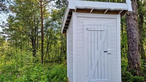 Eine weiße Holzhütte mit einer Tür, umgeben von dichtem Grün und Bäumen.