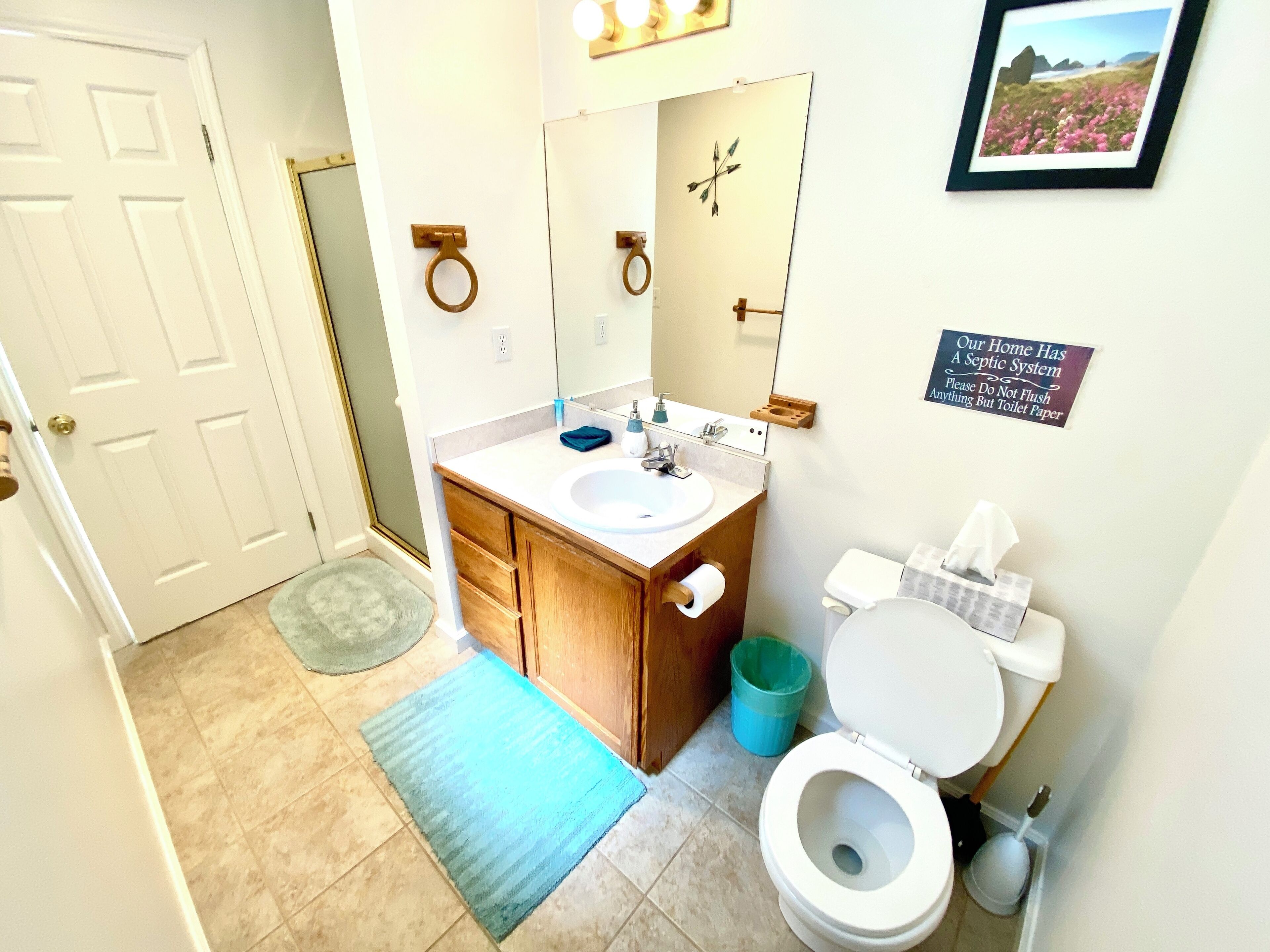 A bathroom with a sink, toilet, and a mirror. There is a sign on the wall and a picture frame on the wall.