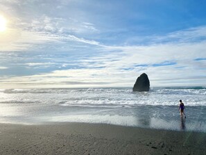 A beach with a large rock formation in the ocean, waves crashing onto the shore, and a person walking in the water.