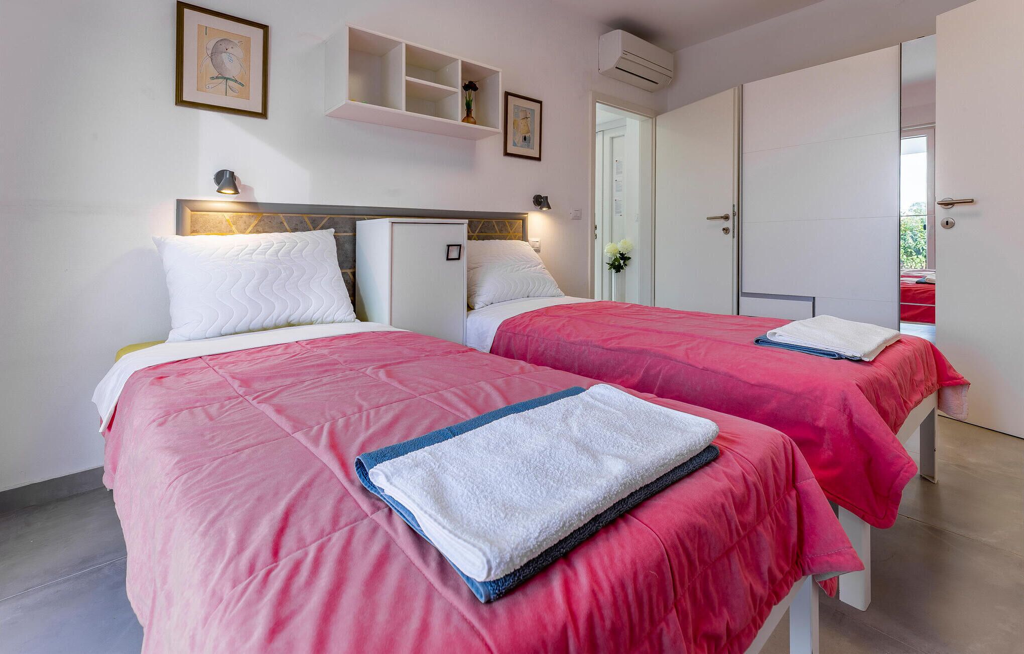 Habitación de hotel con dos camas, cada una con ropa de cama rosa y almohadas blancas. Hay estantes empotrados con objetos decorativos y un cuadro enmarcado.